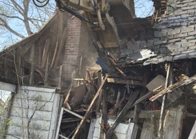 Excavator demolishing a wooden house, with masonry debris and collapsed structure surrounding the site