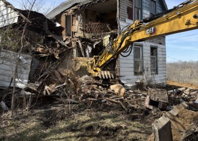 Excavator demolishing an old two-story wooden house, with debris and collapsed structure surrounding the site