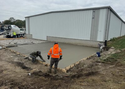 Workers trowel concrete on a large concrete pad while in the background a cement truck driver prepares for next poor.